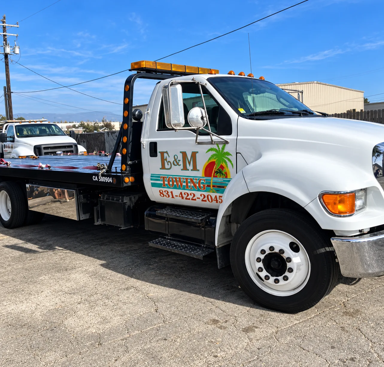 White E&M Towing flatbed truck with a palm-tree logo parked in daylight.