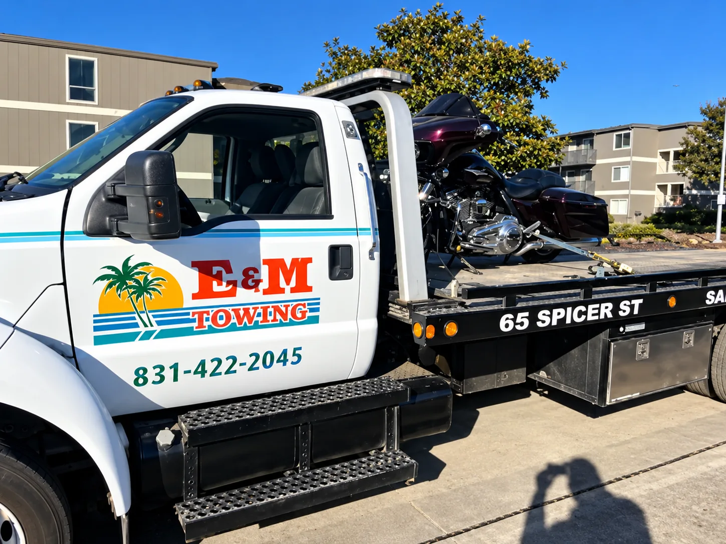 White E&M Towing flatbed truck carrying a motorcycle in daylight, with the business logo and phone number on its door.
